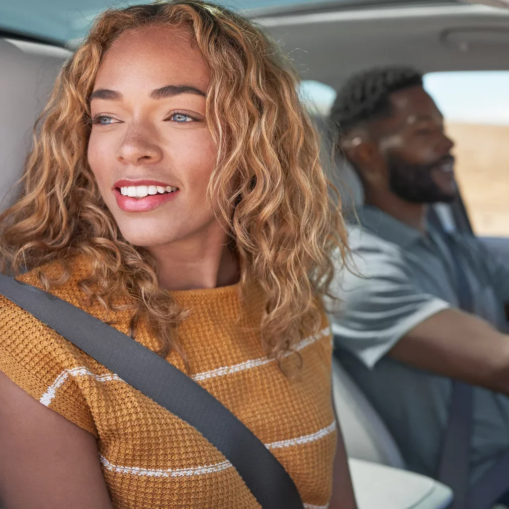 A woman with curly blonde hair and a man in the blurred background drive in a car. The woman is smiling, looking out.